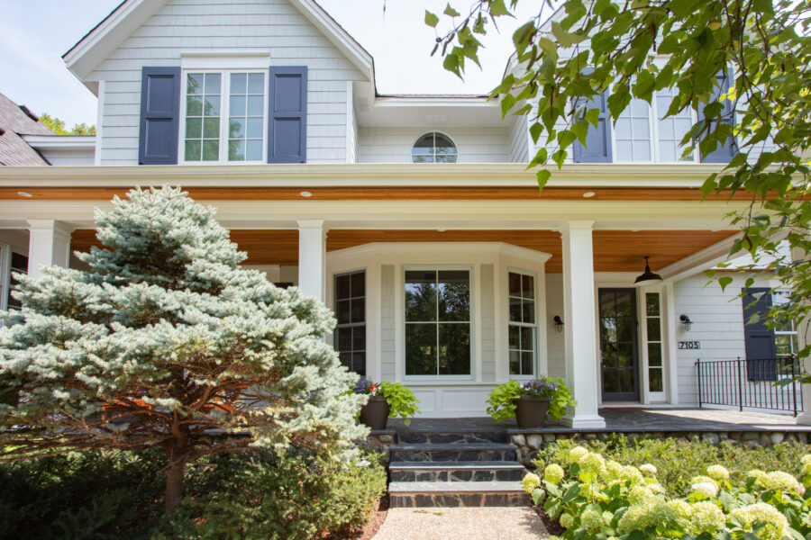 The home exterior features light grey siding, dark blue shutters, and a grand front porch with wood ceilings and slate steps.