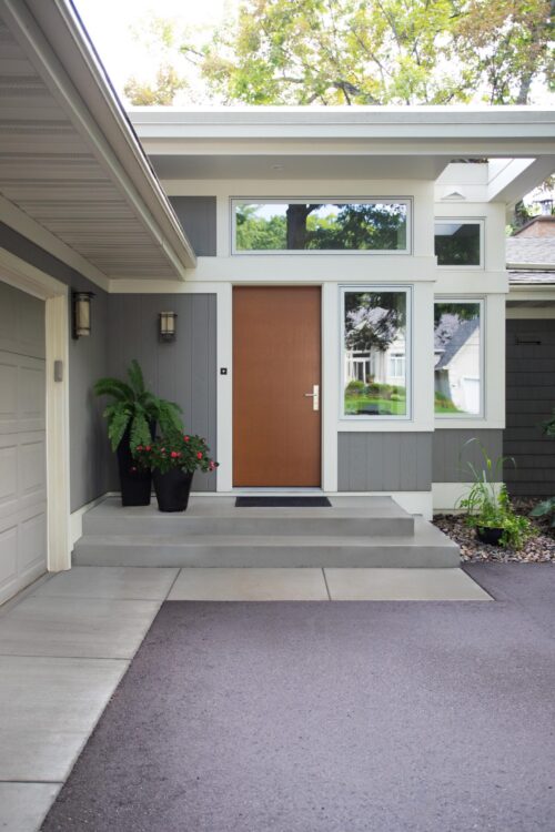 Modern design-build entryway features dark grey siding, warm wood door, large windows, concrete steps.