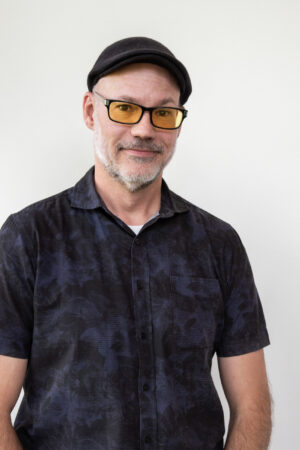 Gray-bearded man smiles, wears black flat cap, yellow glasses, dark patterned short-sleeved shirt on white.