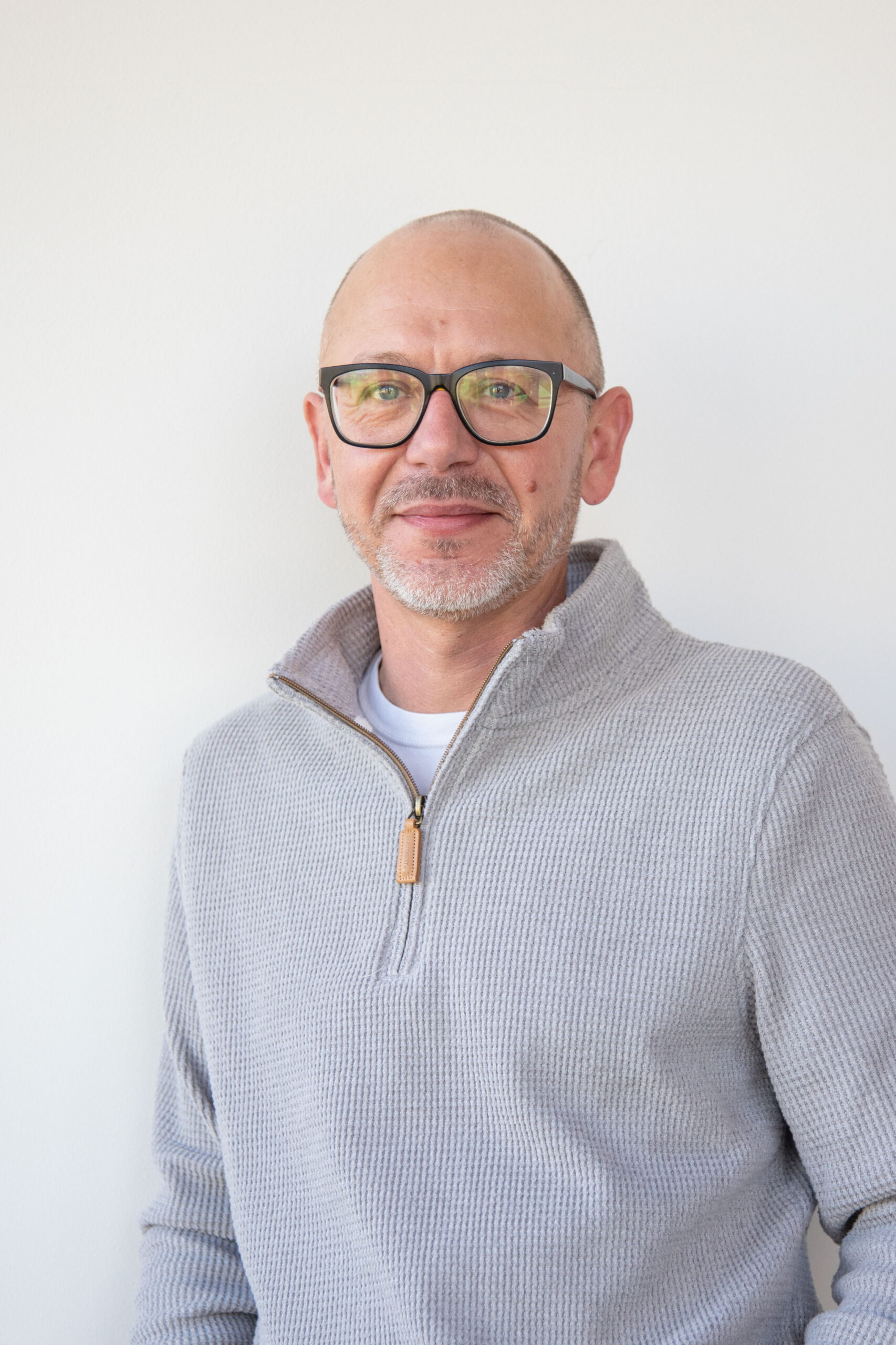 Bald man with beard, dark glasses, textured grey quarter-zip sweater, smiles against white wall.