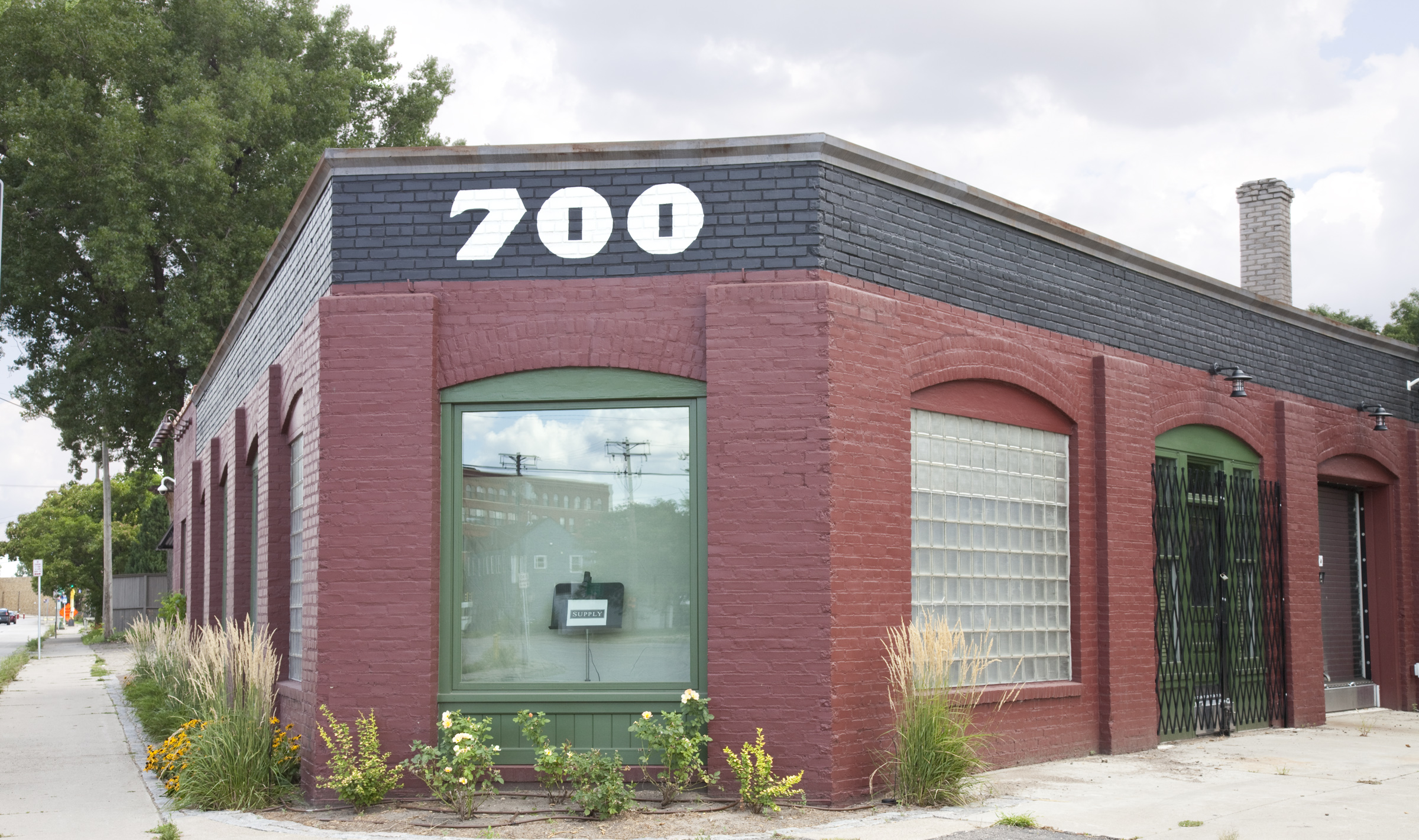 Red brick corner building with large number 200 on dark roof, featuring green-framed storefront windows and arched doorway entrance.