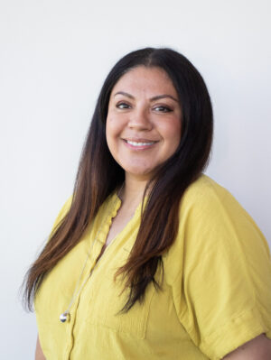 Portrait of a smiling woman with long dark hair wearing a yellow blouse against a light background.