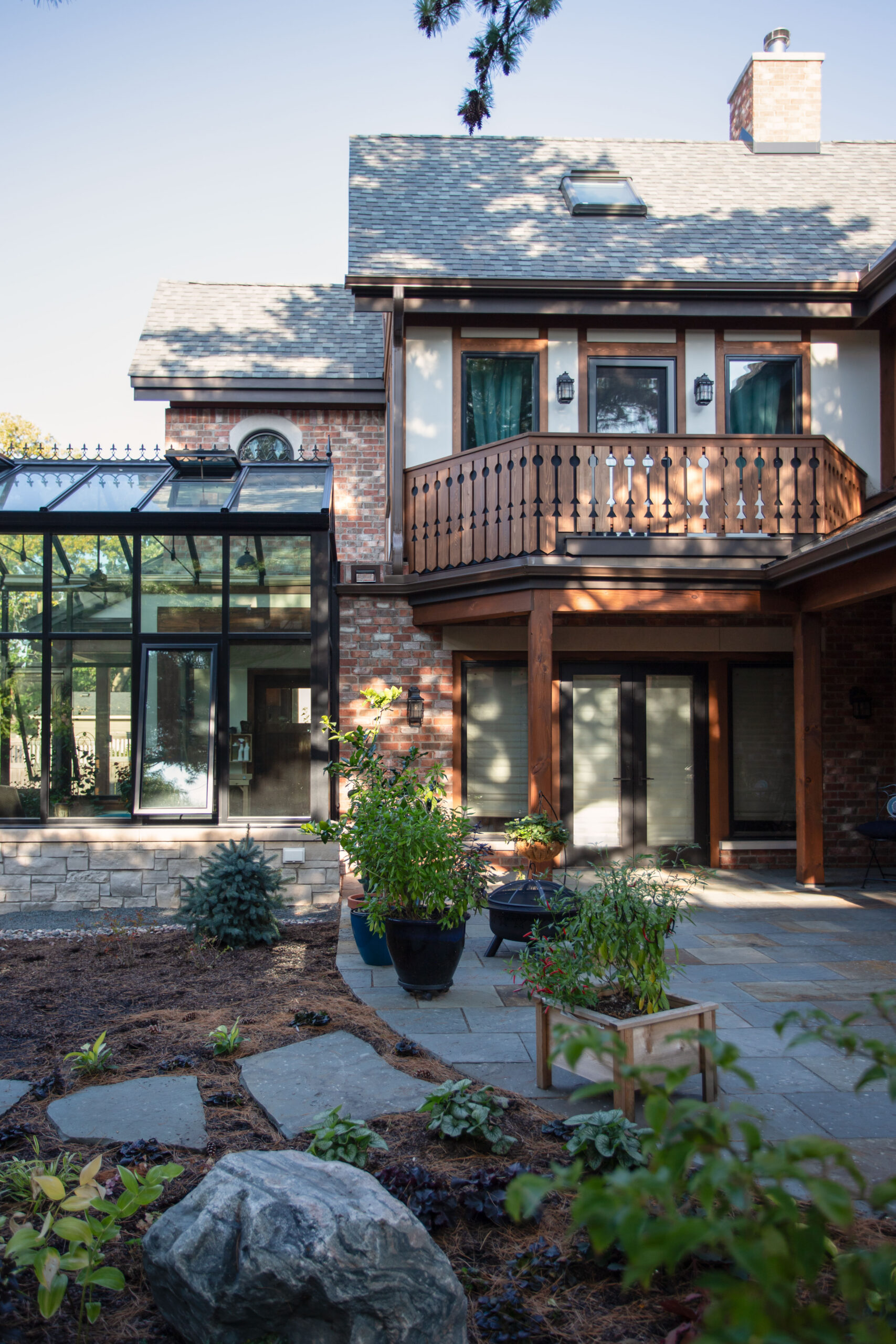 Modern two-story brick house with a wooden balcony and glass conservatory, set in a landscaped garden with potted plants and rocks.