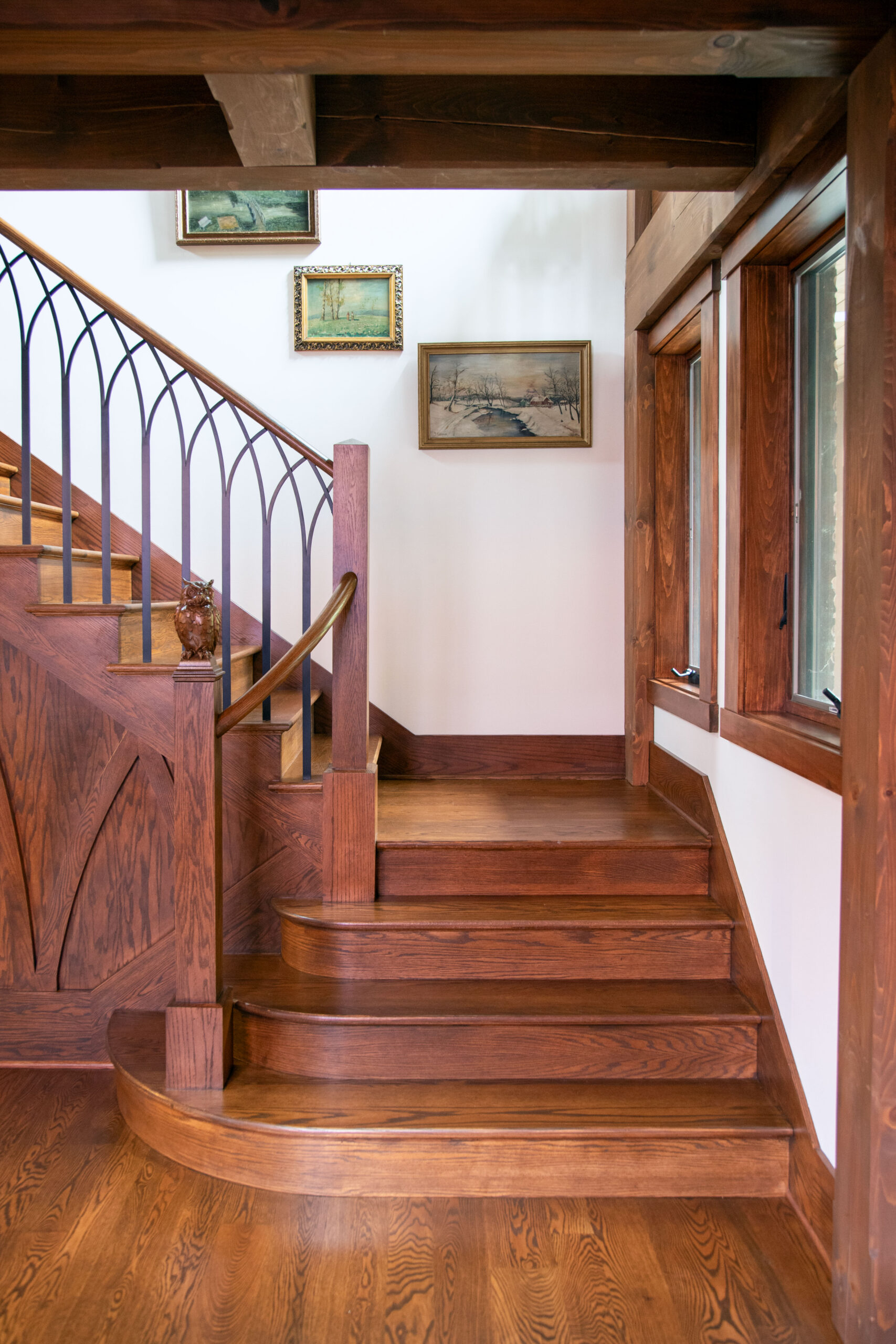 Wooden staircase with ornate iron railing in traditional interior, framed artwork on white walls, exposed beams overhead.