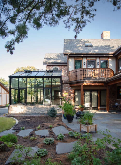 Backyard of a brick house with a glass conservatory, wooden balcony, and potted plants along a stone patio.