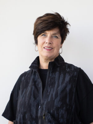 Portrait of a middle-aged woman with short dark hair wearing a black patterned jacket against a light wall.