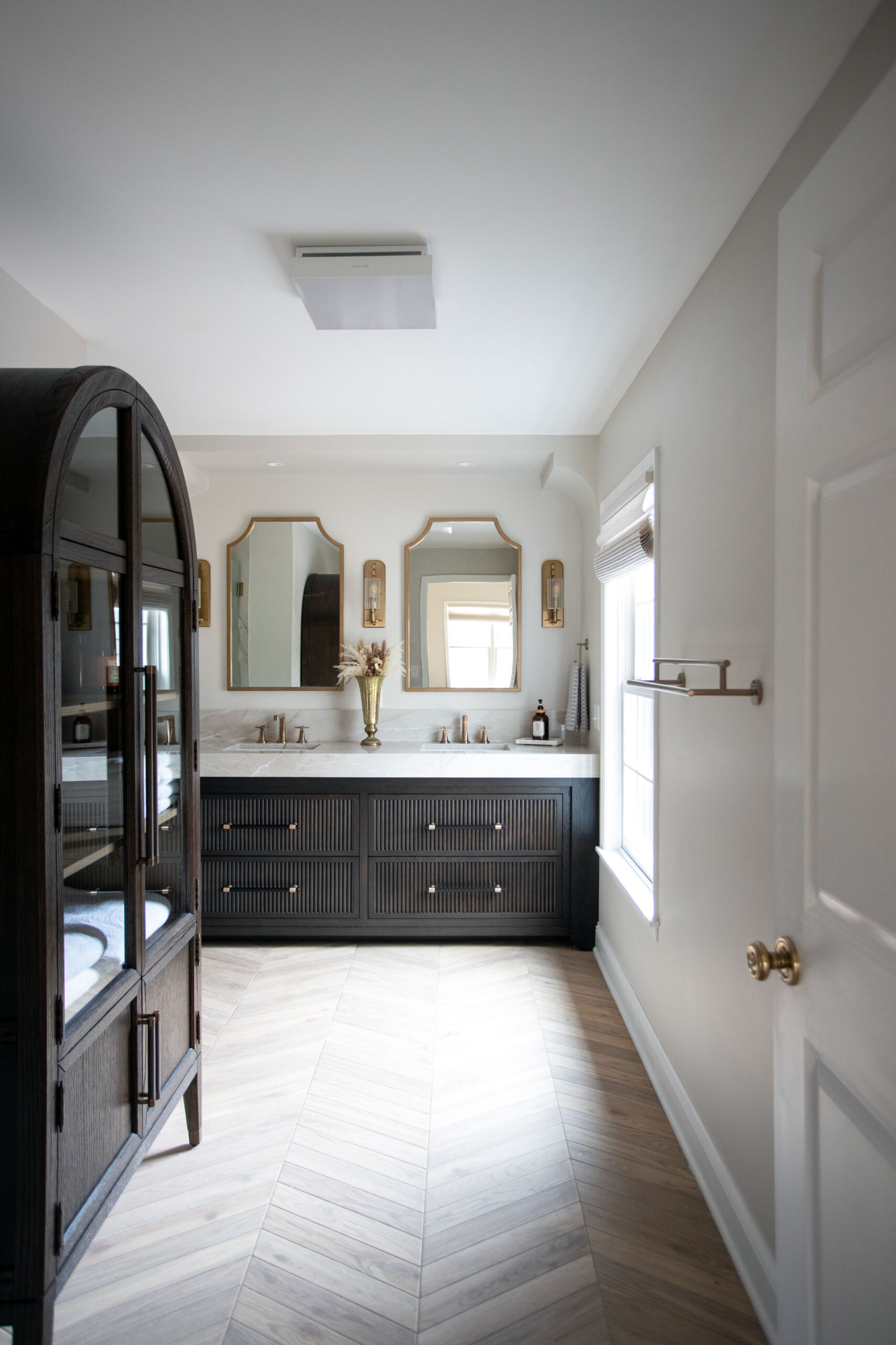 Modern bathroom with black vanity, dual mirrors, arched black door, and herringbone tile flooring