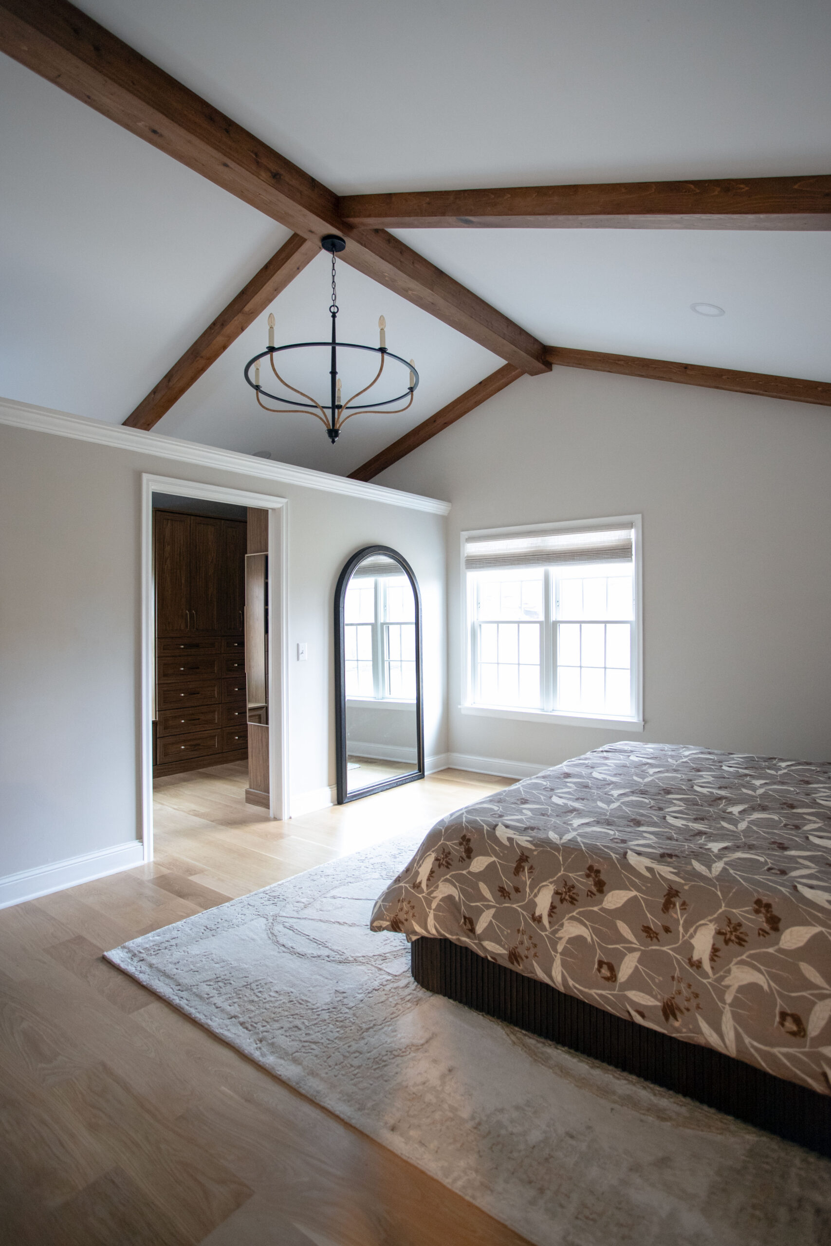 Bedroom with exposed wooden ceiling beams, pendant light fixture, bed with patterned bedding, and windows with natural light