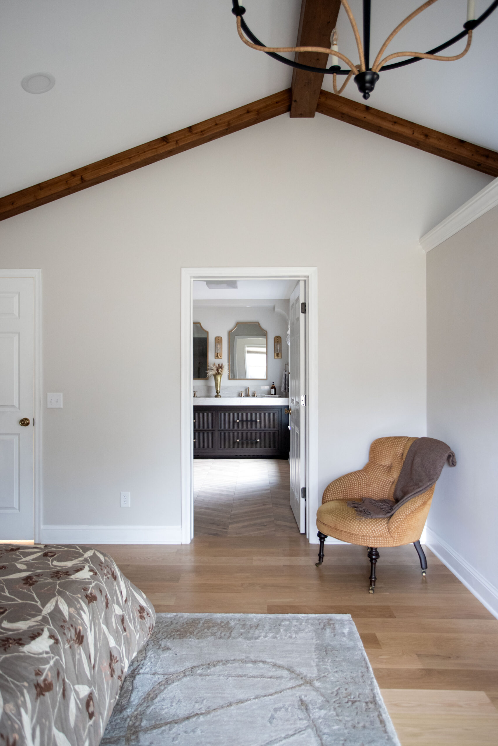 Modern bedroom with exposed wood beams, white walls, hardwood flooring, woven accent chair, and open doorway to ensuite bathroom
