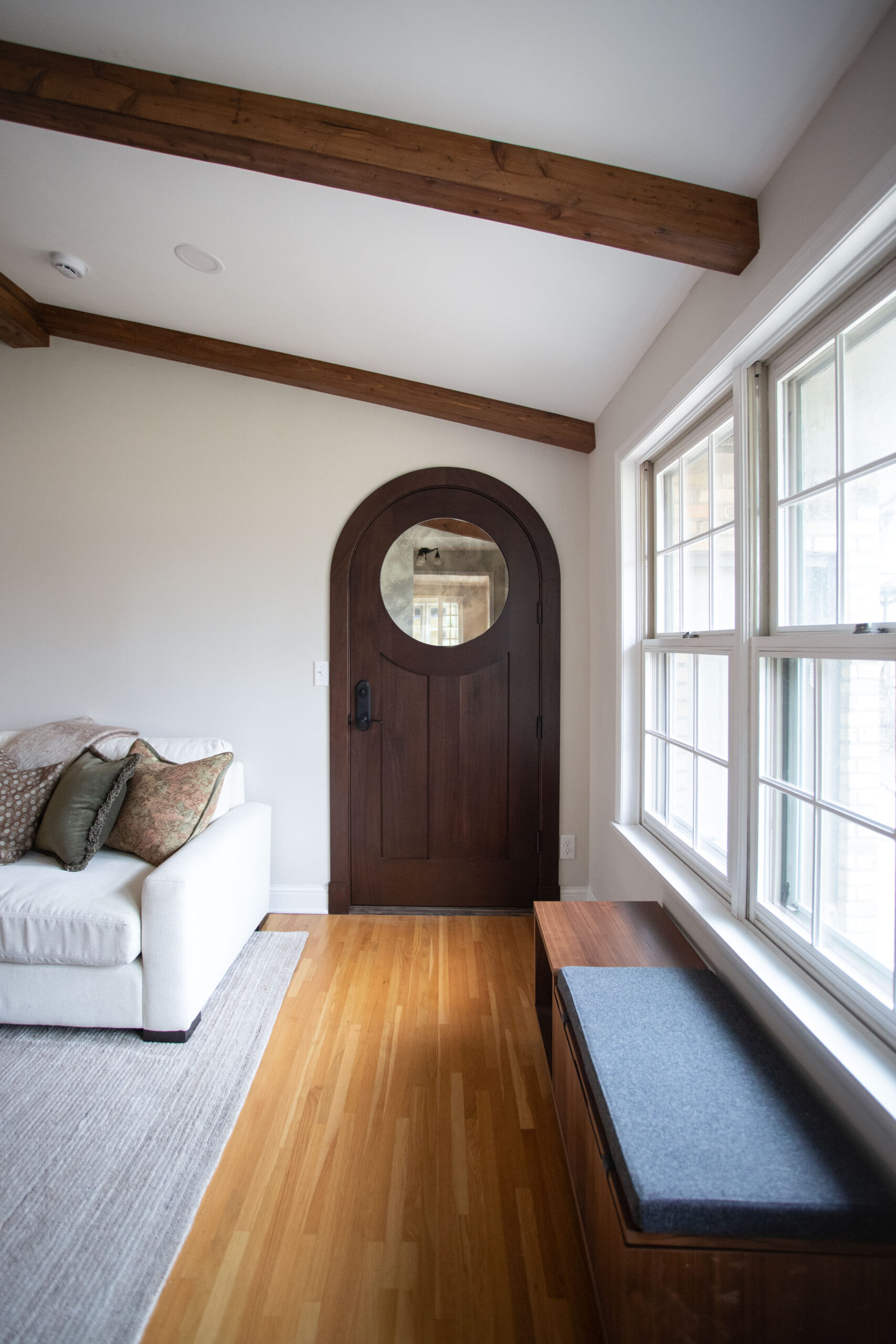 Modern room with arched wooden door, exposed beams, white armchair, and large window seating area