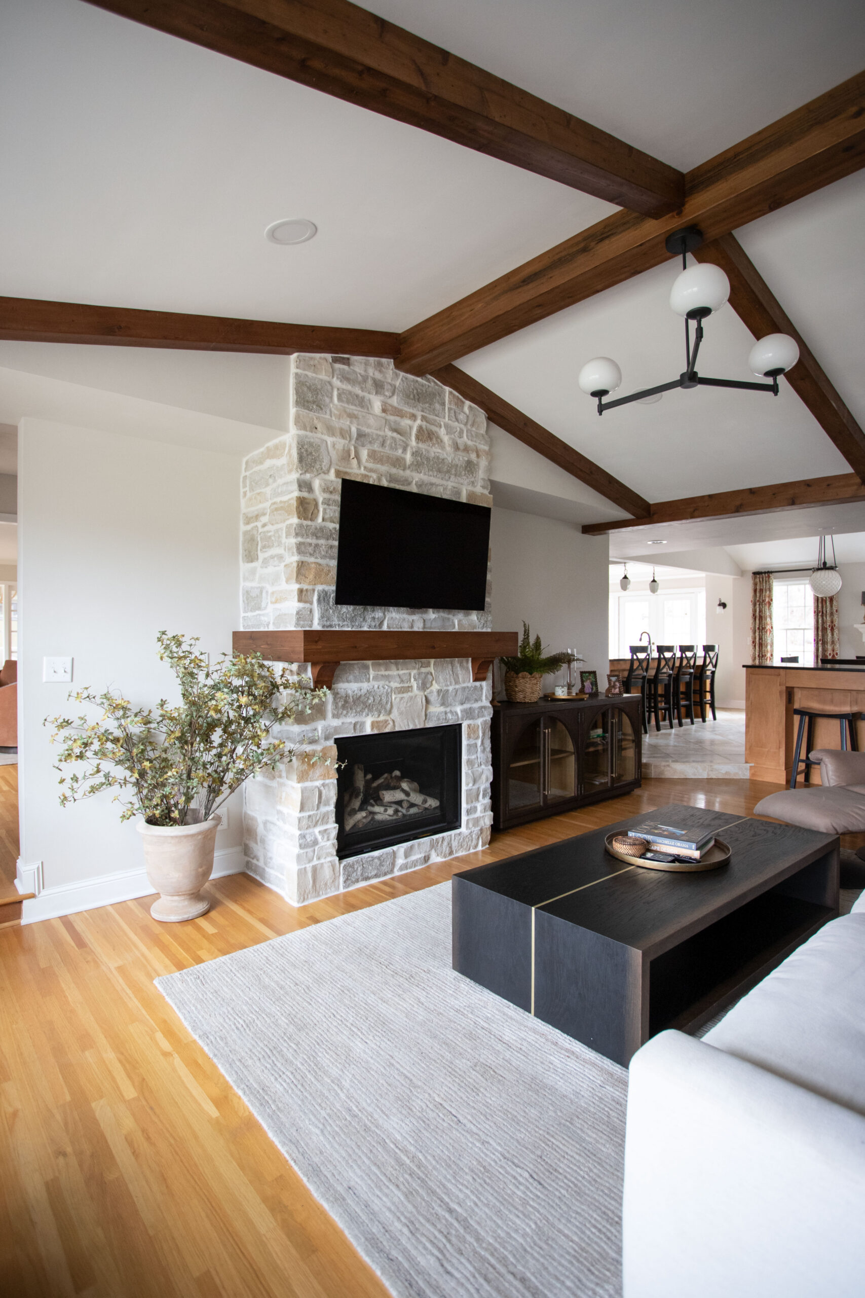 Modern living room with stone fireplace, exposed wood beams, TV above mantel, and dark coffee table on light rug