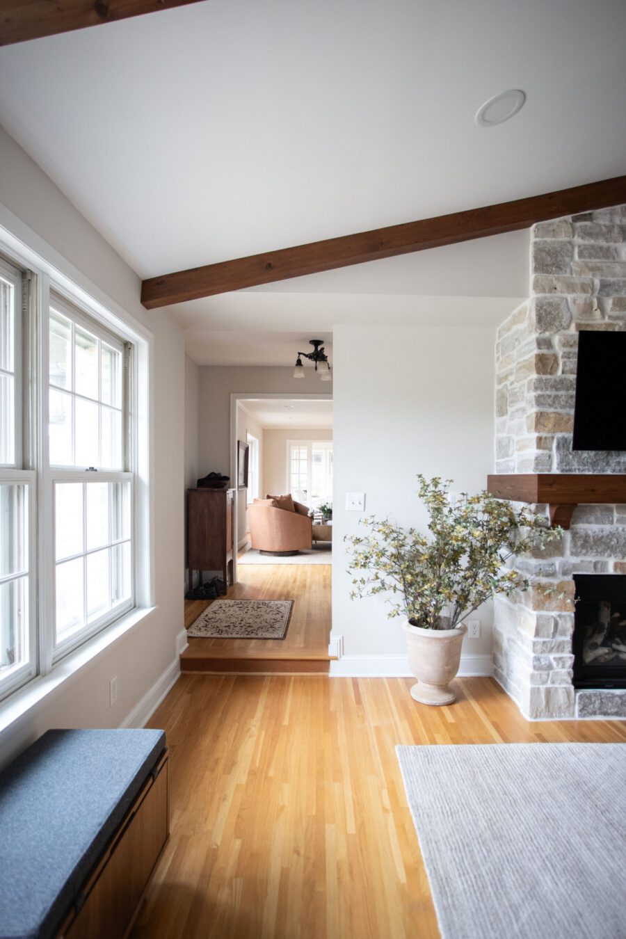 Living room interior with exposed wooden ceiling beams, a stone fireplace on the right, and large windows on the left letting in light.
