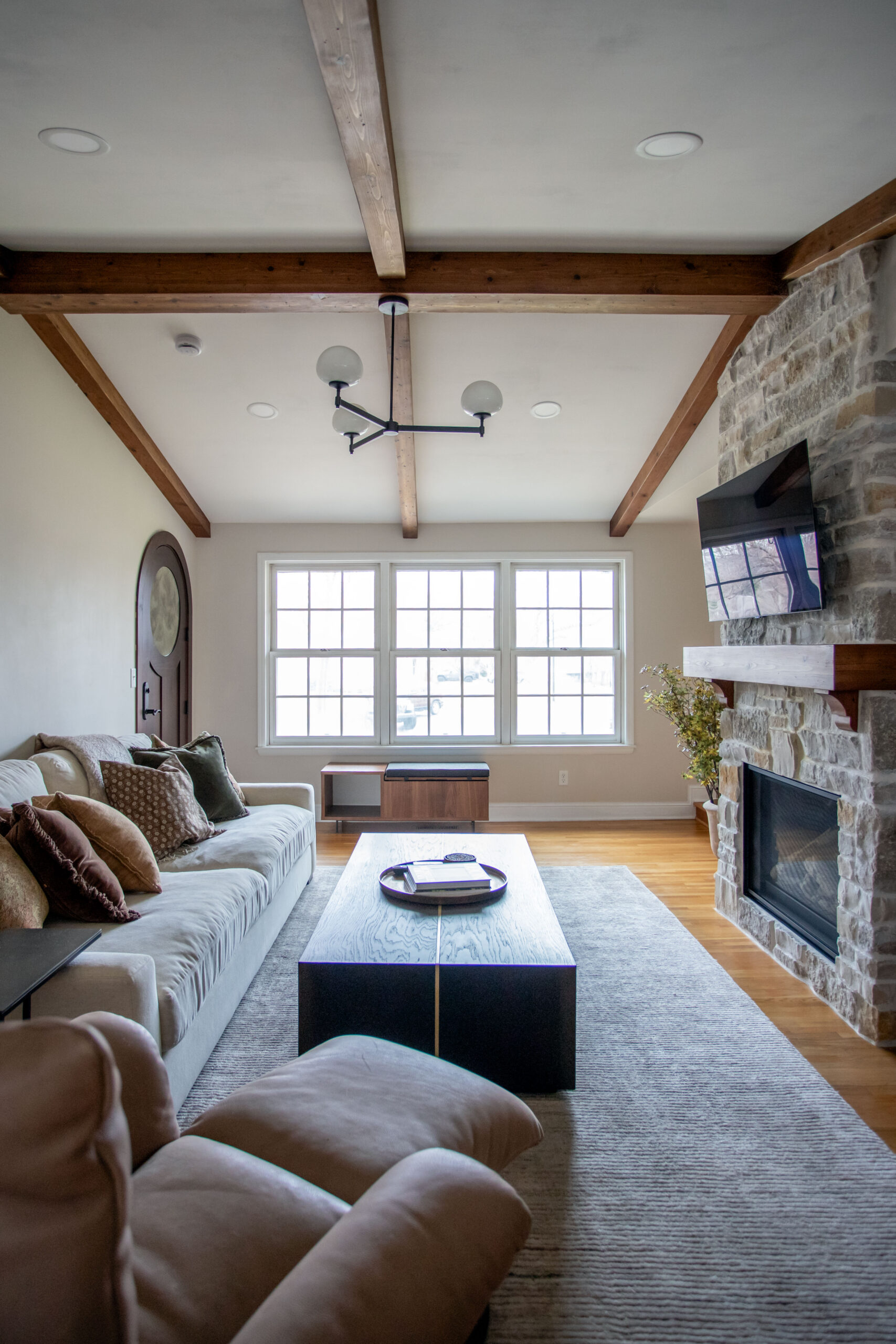 Modern living room with exposed wood beams, stone fireplace, large windows, and contemporary furnishings arranged around a black coffee table.
