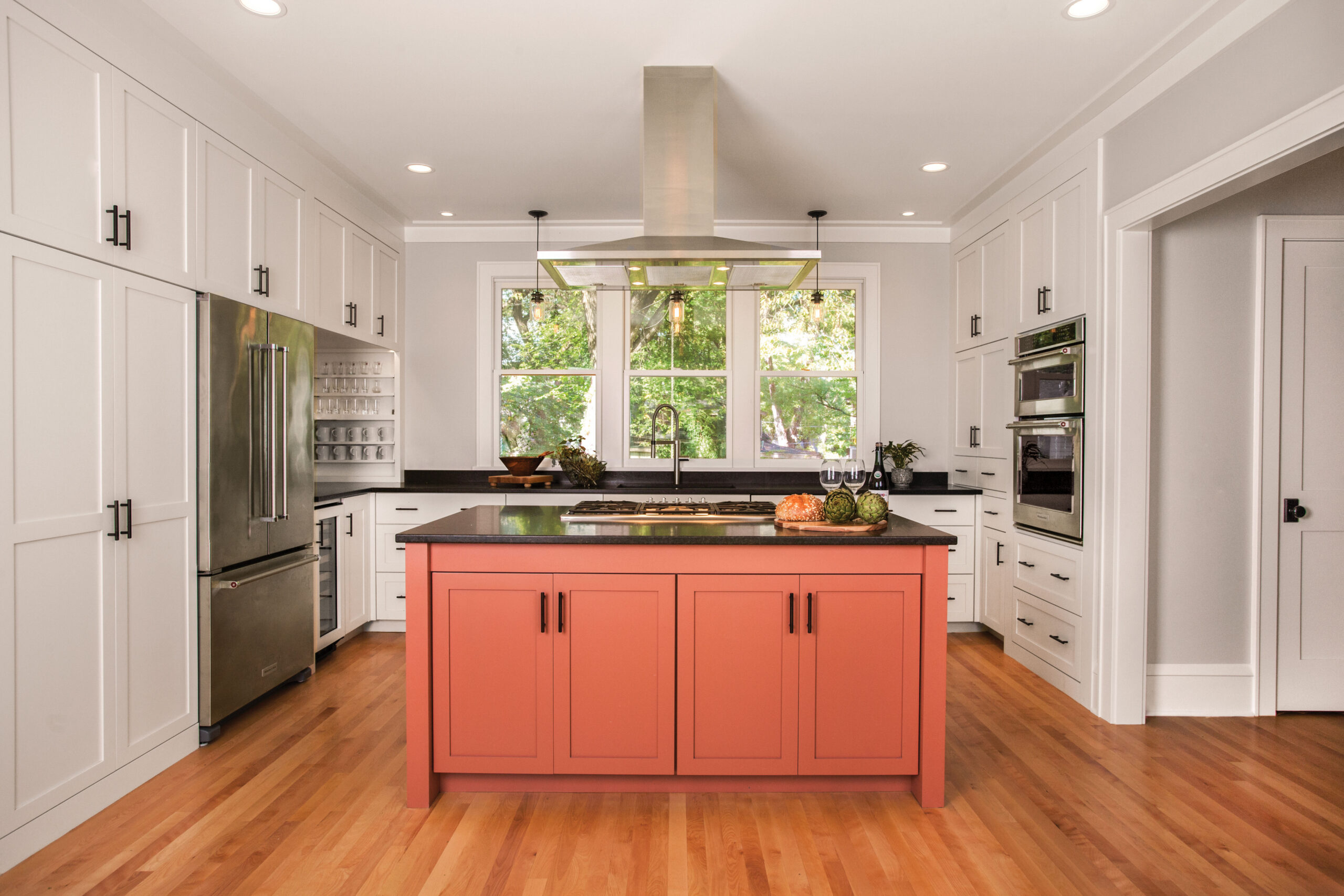 Bright kitchen with white cabinets and a coral pink island, stainless steel appliances, and large windows showing greenery outside