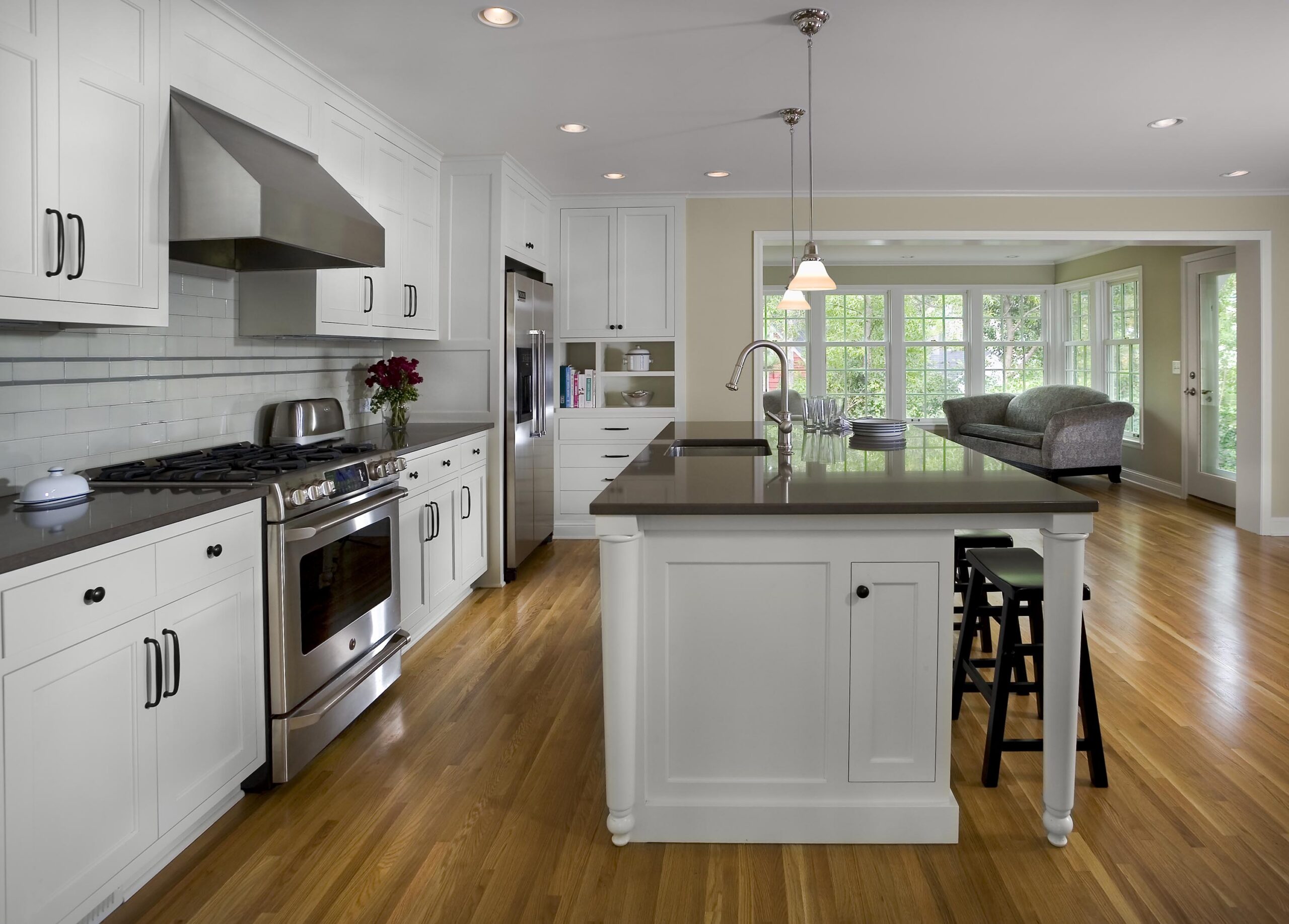 Bright white kitchen with shaker cabinets, stainless steel appliances, and a large island with a sink; opens to a sunlit living area with windows.