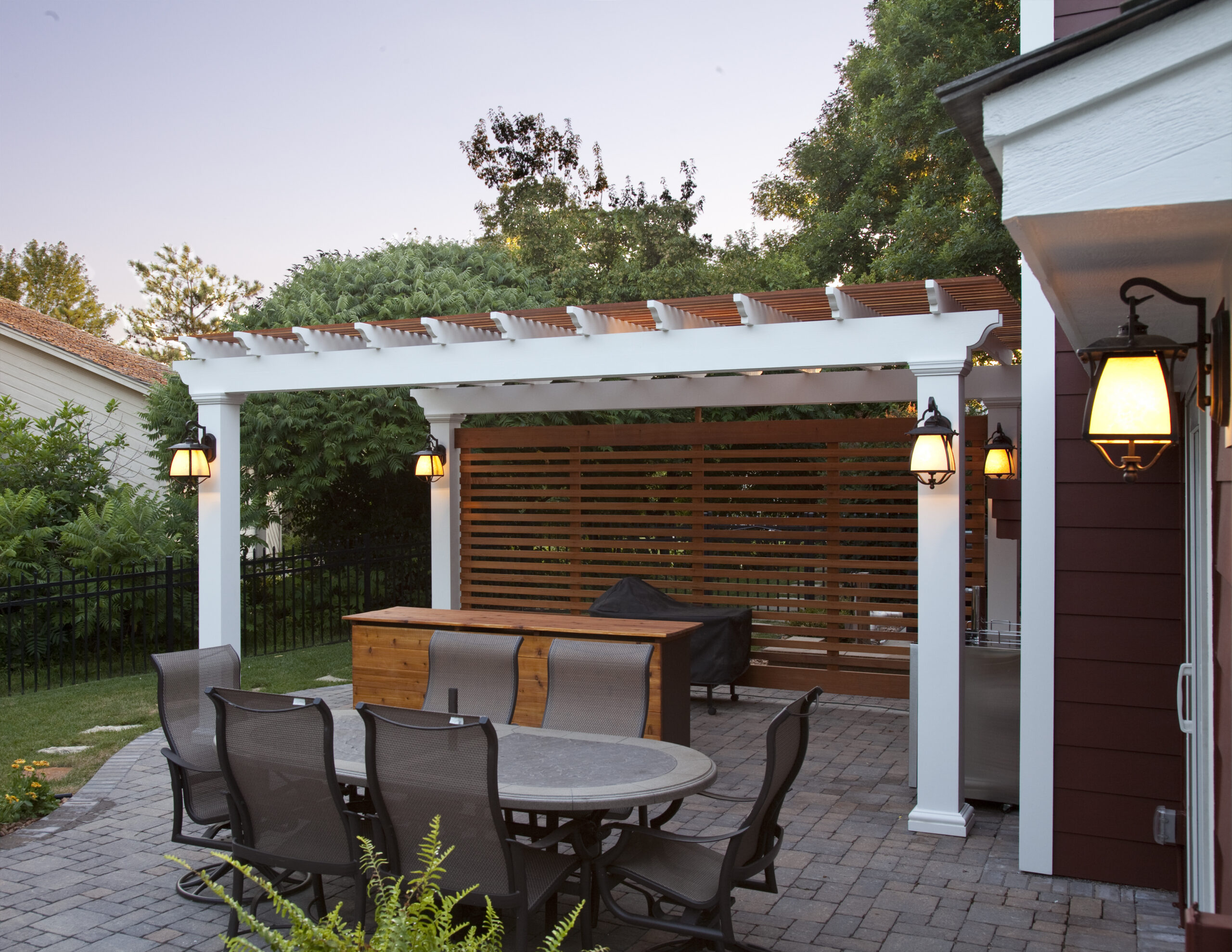 Outdoor patio dining area under a white pergola with a wooden slat privacy screen and warm lantern lighting, surrounded by greenery.