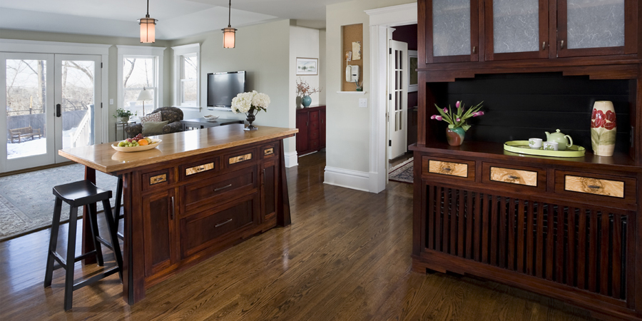 Open-plan kitchen with a dark wood island, bar stool, fruit bowl, and hardwood floors, leading to a living area and French doors.