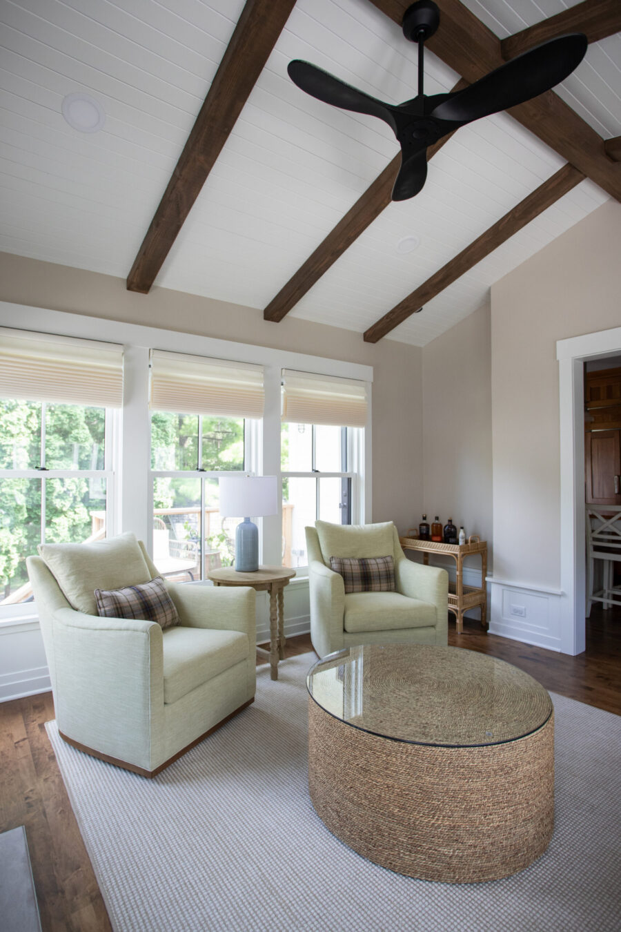 Bright living room with pale green armchairs, a woven round coffee table, and a wicker side table near large windows.