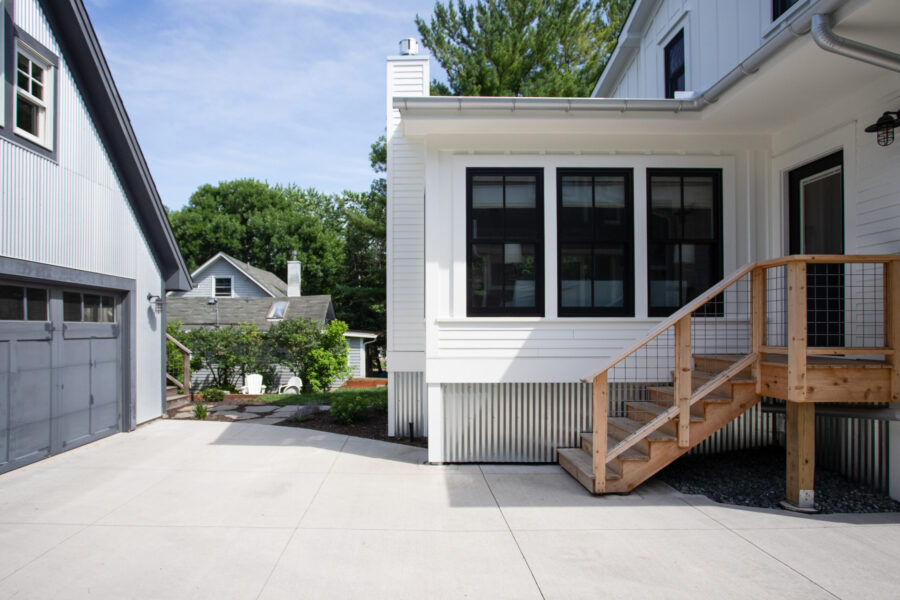 White exterior of a modern house with large black-framed windows and a wooden staircase on a concrete patio