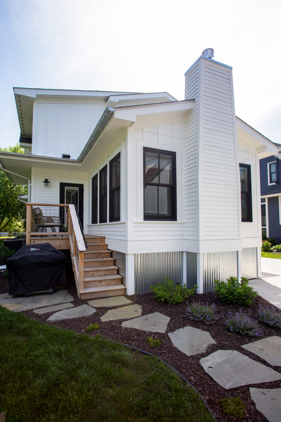 White modern house exterior with a tall white chimney, large black-framed windows, and a wooden staircase leading to a small porch area with seating.