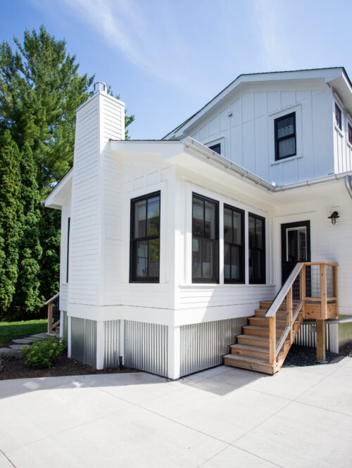 White two-story house with black-framed windows, a tall white chimney, and wooden stairs to a raised entry under a blue sky.