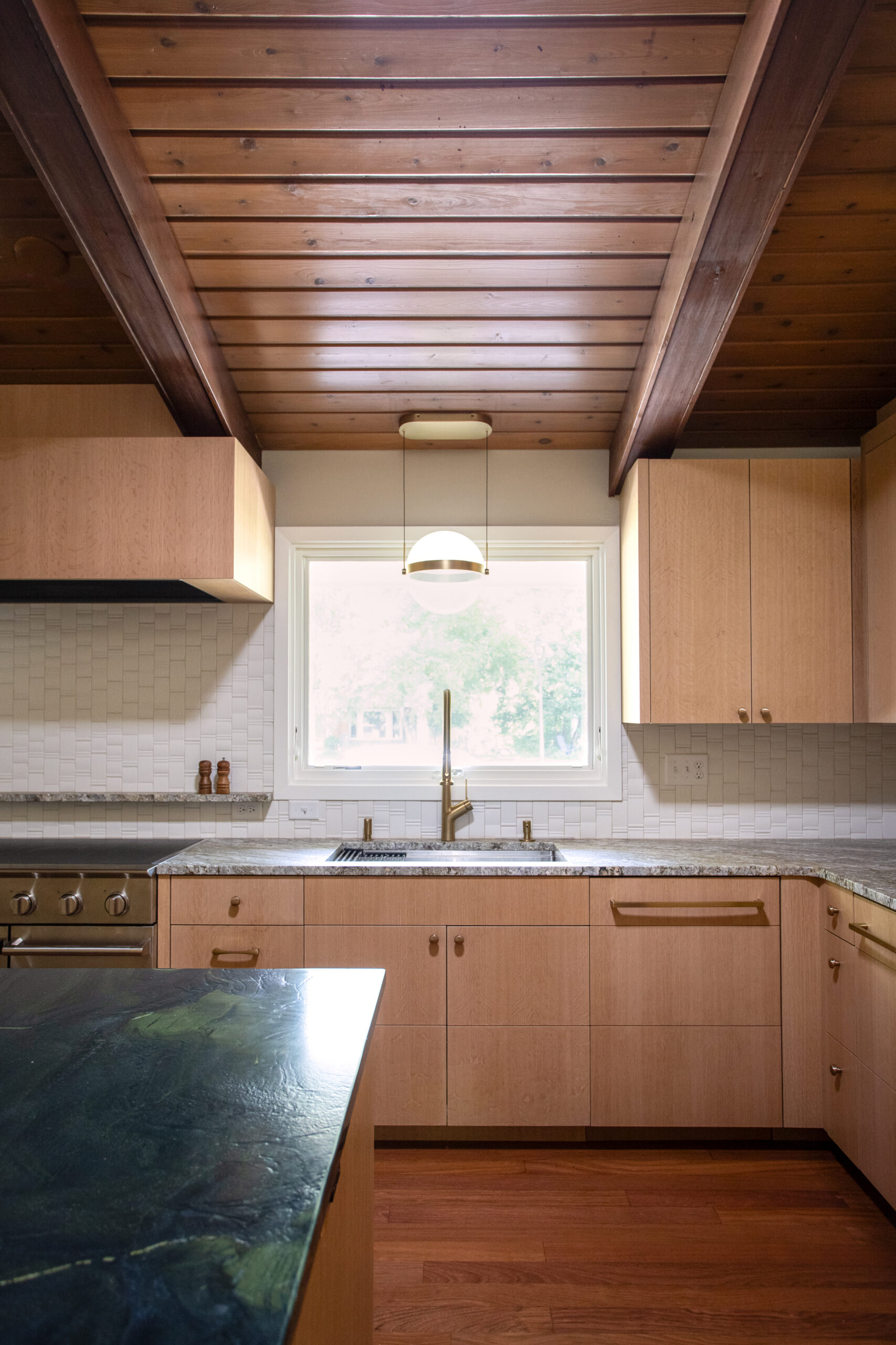 Wood-paneled ceiling with exposed beams over a modern kitchen, light maple cabinets, and a window above the sink.