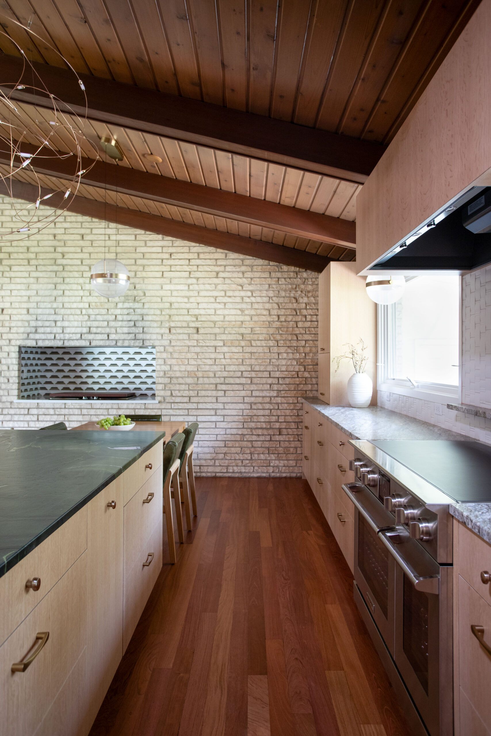 Modern kitchen with brick wall, wood ceiling beams, and stainless steel range on the right; green-topped island on left with bar stools.