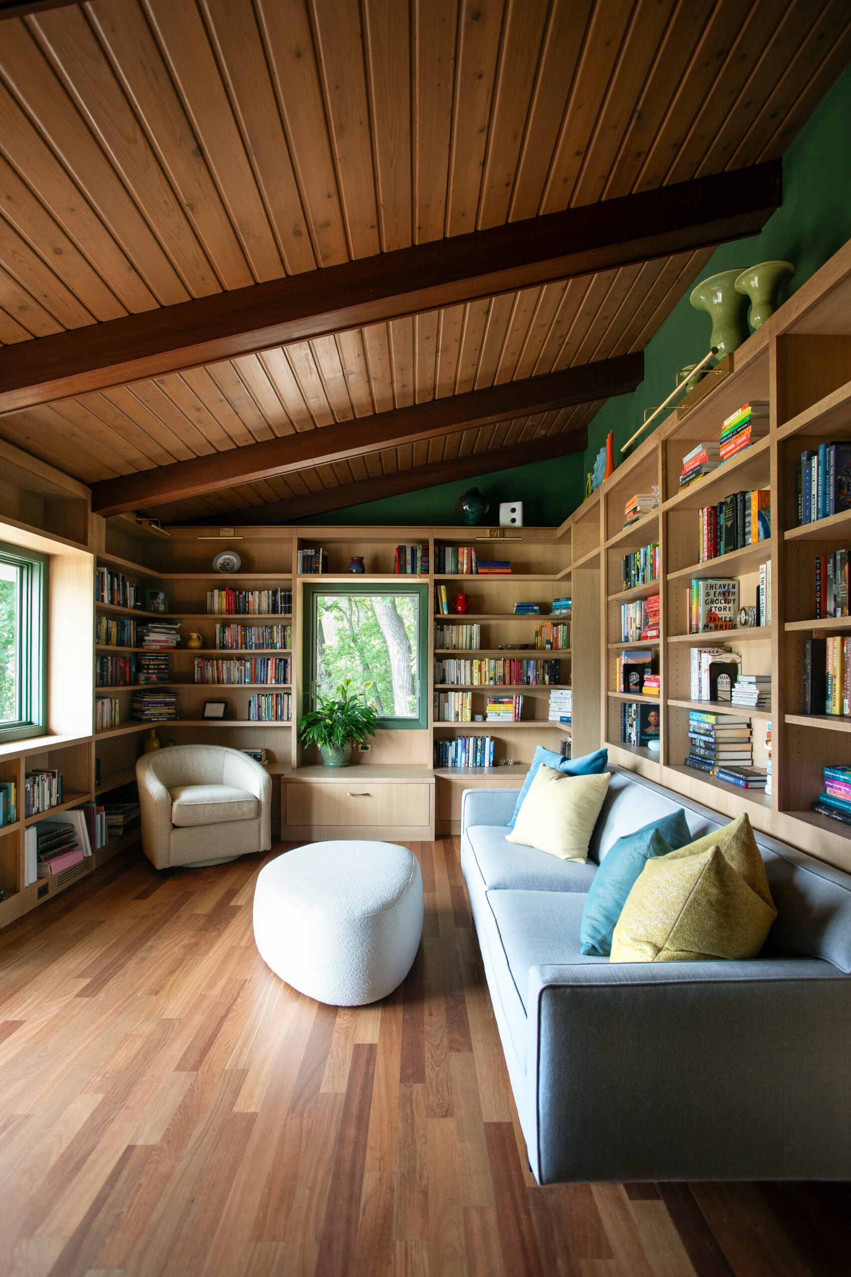 Cozy living room with wooden vaulted ceiling, built-in book shelves, and a light gray sectional with colorful pillows facing a round white ottoman.