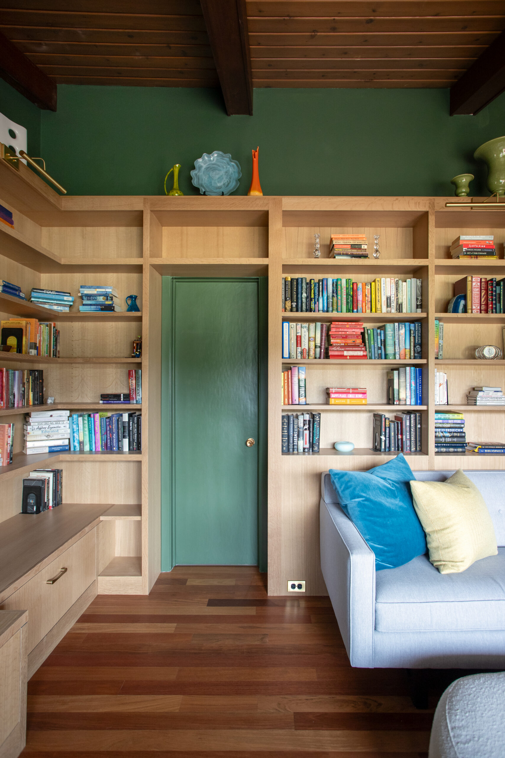 Built-in wooden bookshelves surround a green door on a green-walled wall, with decorative vases on the top shelf and a blue sofa with colorful cushions nearby.