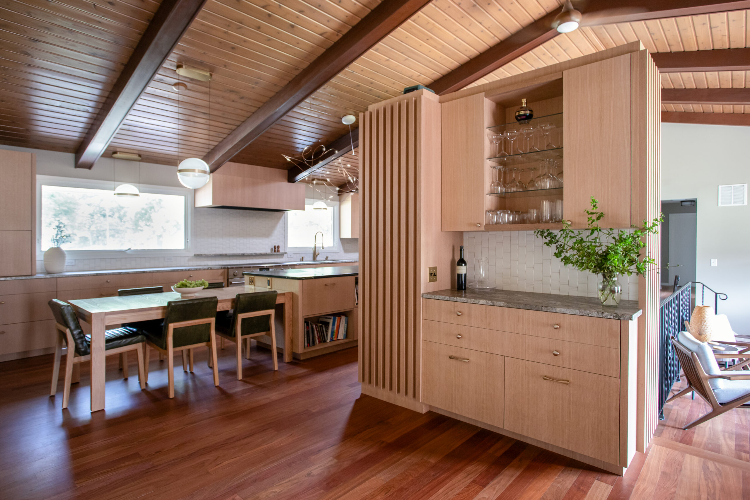 Open-concept kitchen with wood cabinets, a glass-front display, and a wooden dining table under a vaulted, wood-paneled ceiling.