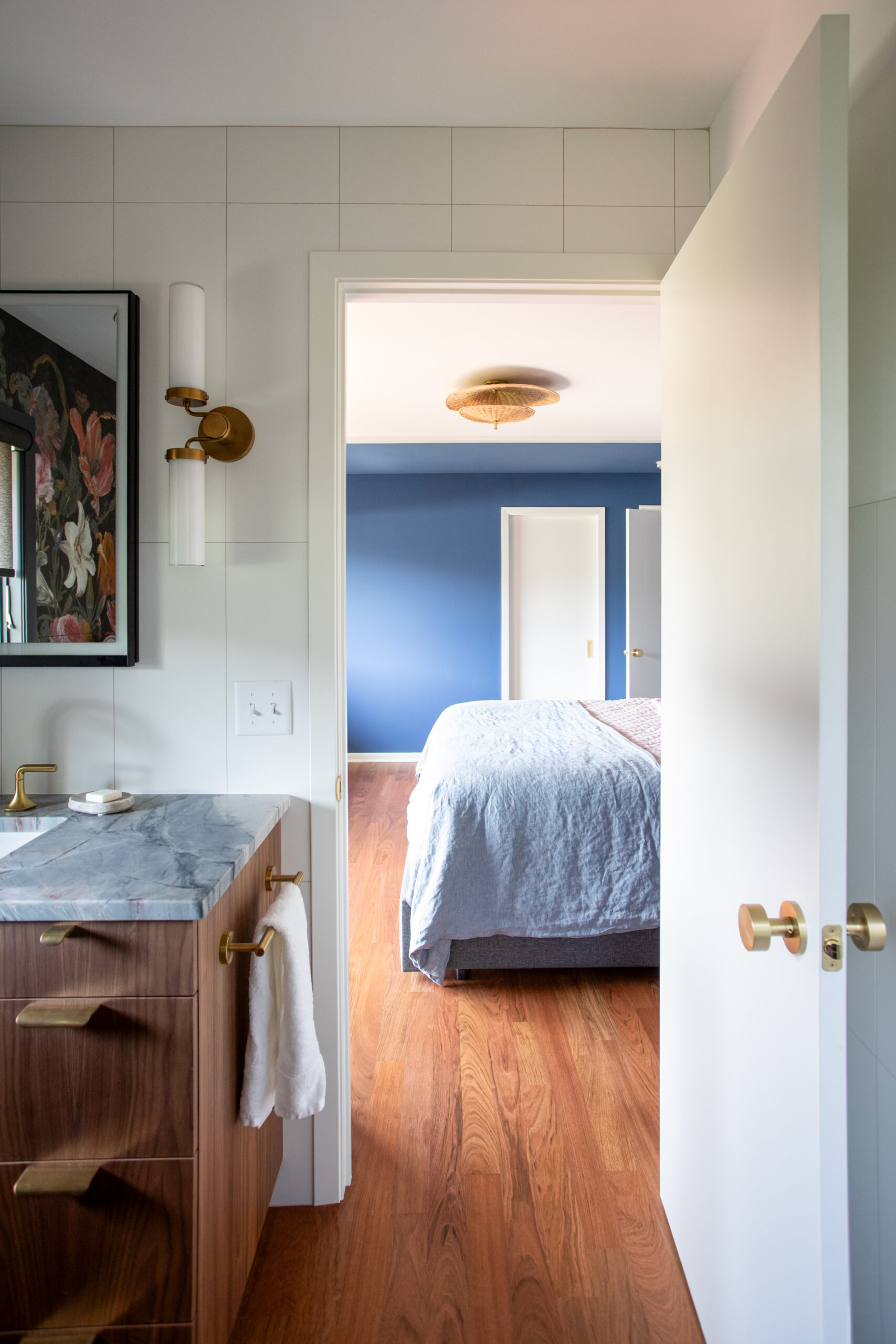 Bathroom vanity with marble countertop and brass fixtures; towel hangs from the side, open door to a blue bedroom with a bed visible beyond.