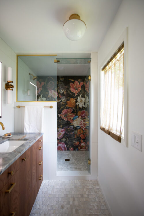 Modern bathroom with white walls, wood vanity, and gold fixtures; glass shower enclosure with floral wallpaper visible behind.