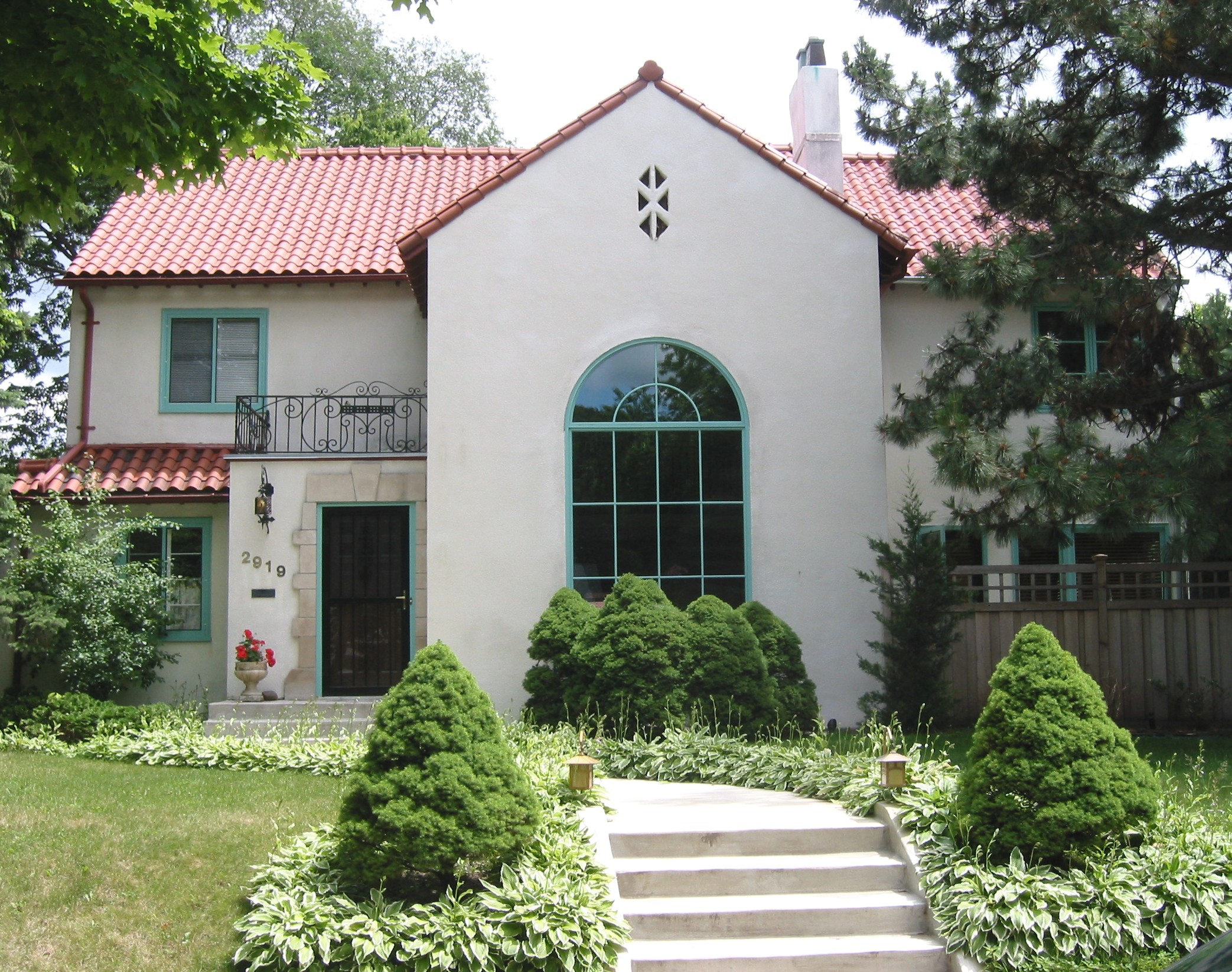White stucco house with teal window frames, red tile roof, and a manicured garden with rounded shrubs in the foreground.