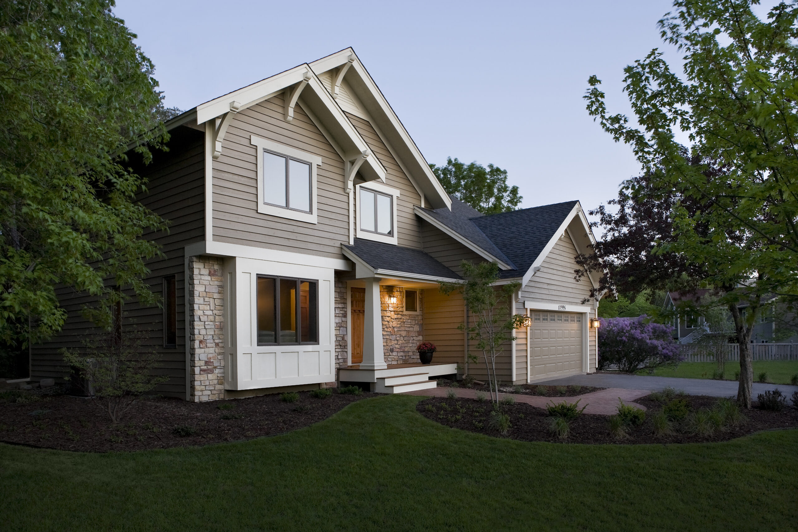 Front view of a two-story Craftsman-style house with beige siding, stone accents, white trim, and a two-car garage, surrounded by green trees at dusk/early evening.