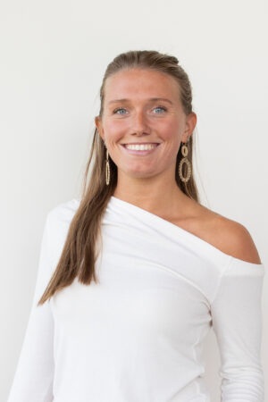 Smiling woman with long hair in a white off-the-shoulder top and gold dangling earrings against a light background.