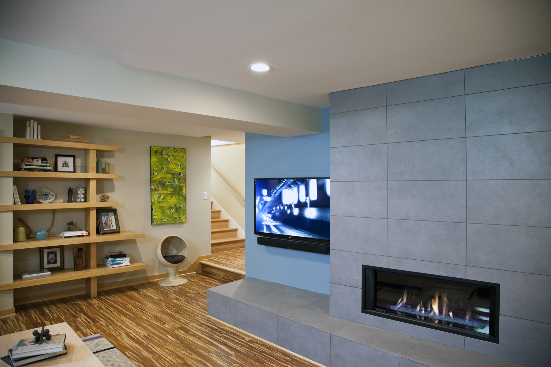 Modern living room with a gray tiled fireplace surround, wall-mounted TV, and open wooden shelving displaying books and decor.