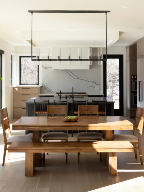 Modern kitchen with a large wooden dining table and benches, marble backsplash, and a suspended rectangular light fixture over the island.
