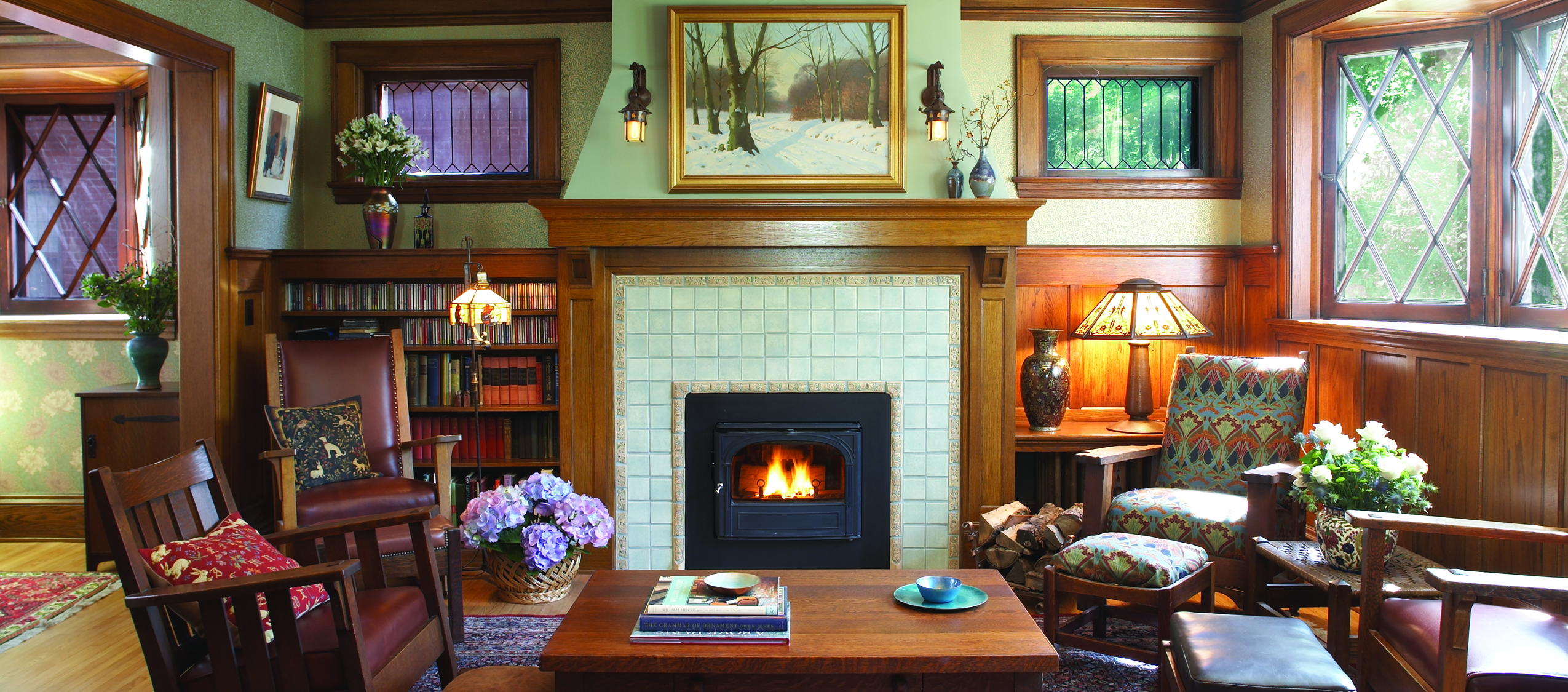 Cozy living room with a lit tiled fireplace, wooden mantel, and green walls framed by dark wood trim. Built-in bookshelves line the left wall.