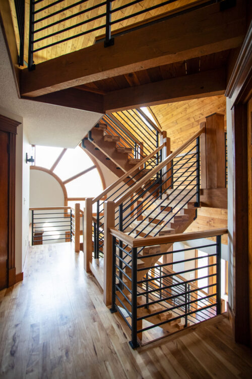 Wooden staircase with black metal railing in a bright multi-level interior with warm wood paneling and a large arched window letting in light.