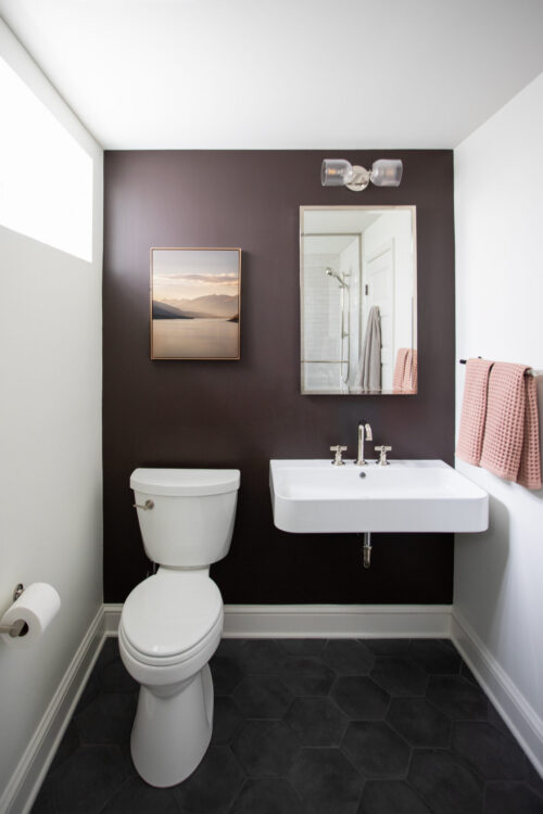 Modern bathroom with a dark accent wall, white sink, and toilet with pink towels on the right mirrored by a rectangular mirror above the sink