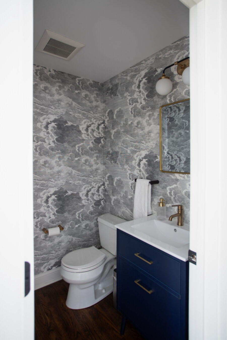 Narrow bathroom with a dark blue vanity, white sink, brass fixtures, and cloud-pattern gray wallpaper.