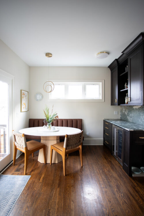 Dining nook with a round white table, brown bench seating, and wooden chairs in a bright kitchen corner.