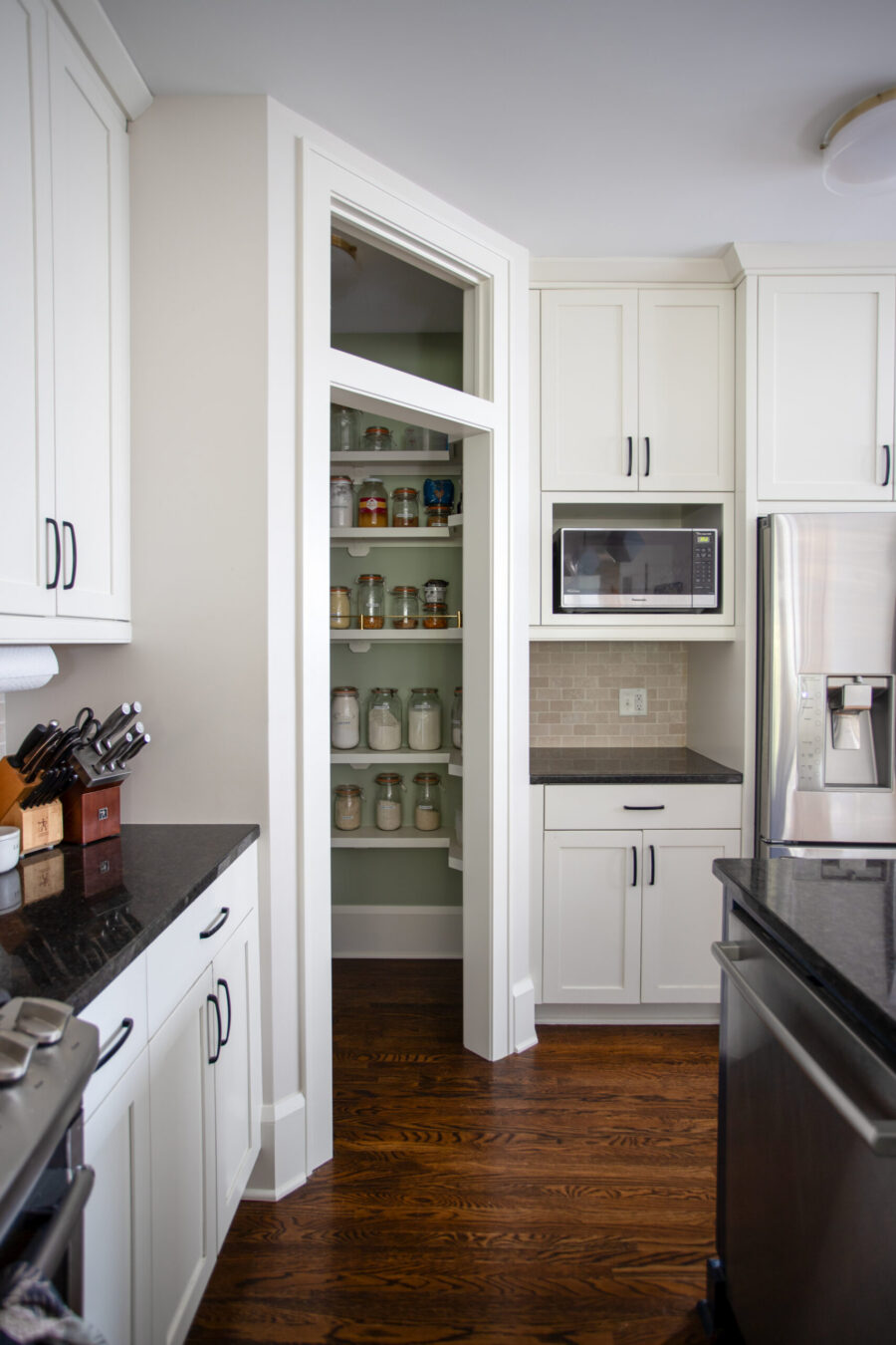 Open pantry with glass jars and mason jars inside, surrounded by white cabinets in a bright kitchen.