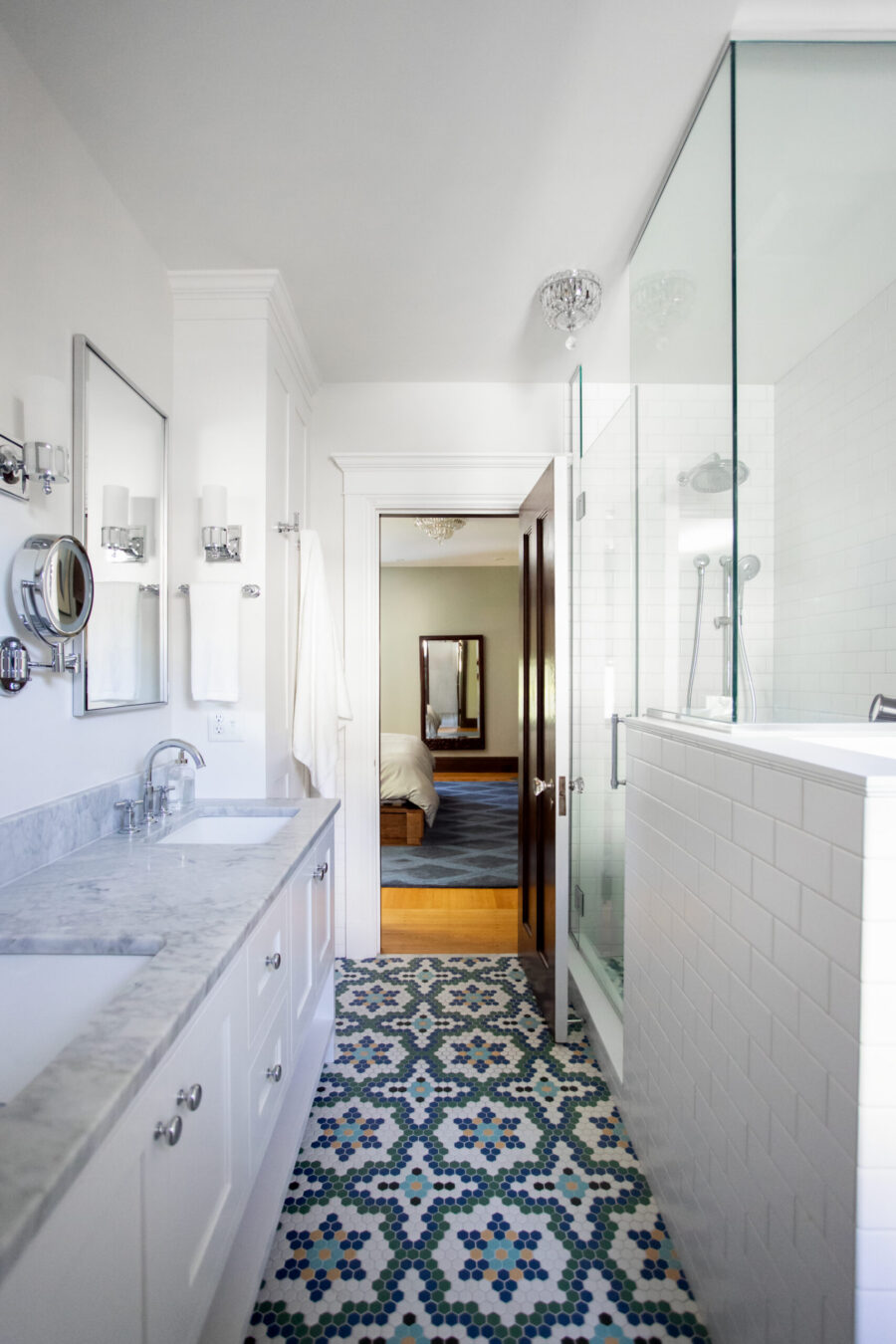 White, modern bathroom with marble counter, double sinks, and wall-mounted lights; glass shower enclosure on the right, doorway opening to a bedroom beyond.