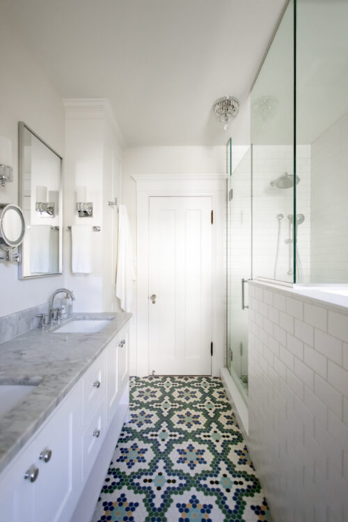 Bright white bathroom with a patterned blue-green floor, double sink vanity, and glass shower enclosure.