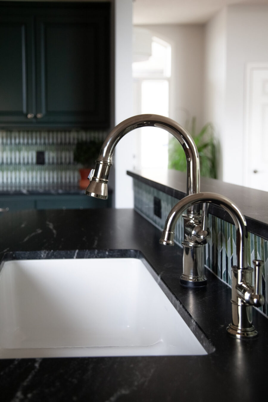 Close-up of a shiny chrome kitchen faucet over a white sink, with a black granite countertop and a green tile backsplash in the background.
