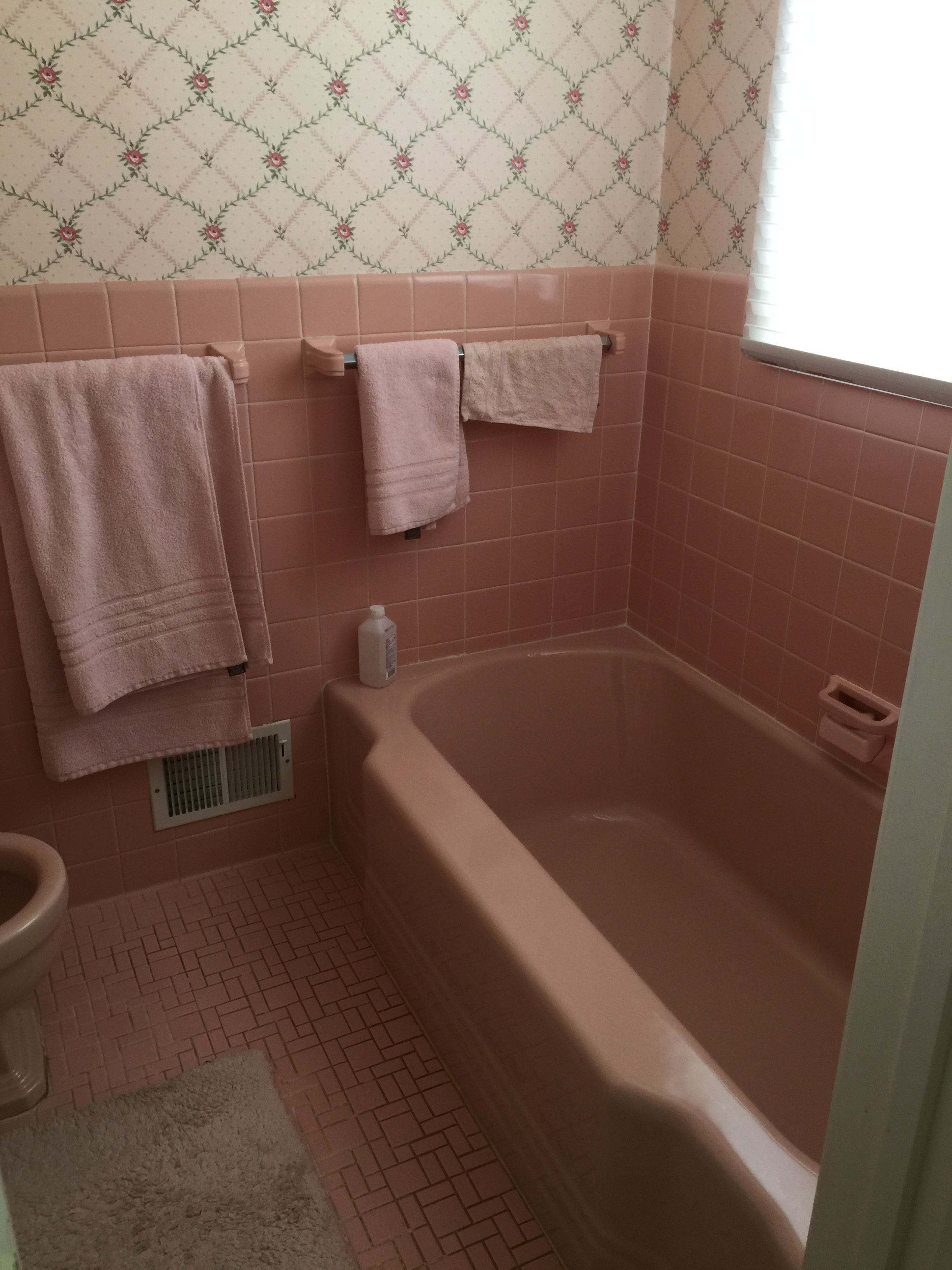 Vintage pink bathroom: pink-tiled walls and tub, pastel towels on rack, floral wallpaper, window with blinds.