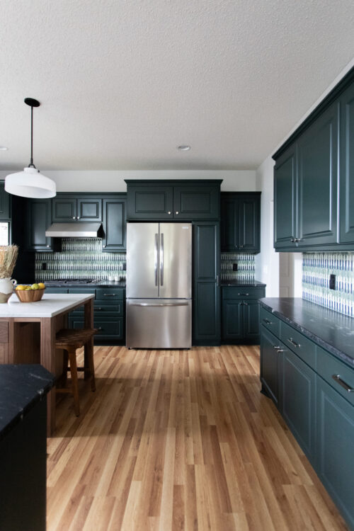Modern kitchen with dark teal cabinets, stainless steel refrigerator, pendant light, and a wooden island with fruit bowl on a light hardwood floor.